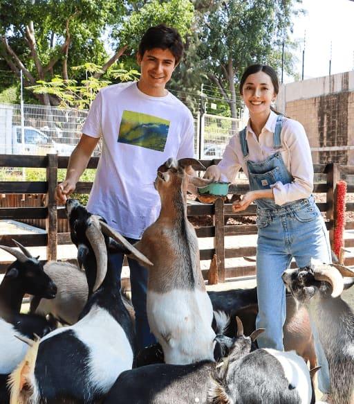 Dos voluntarios jóvenes sonríen mientras alimentan a un grupo de cabras en un corral al aire libre del acuario.